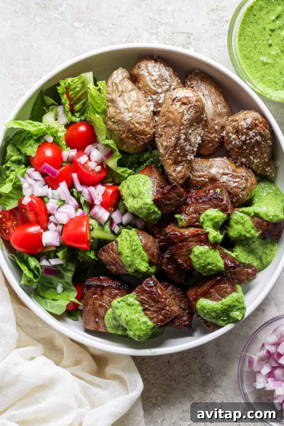 A beautifully presented bowl filled with grilled steak chunks, drizzled with a rich green sauce, accompanied by golden roasted potatoes, and a fresh side salad of crisp lettuce, juicy tomatoes, and finely chopped red onions.