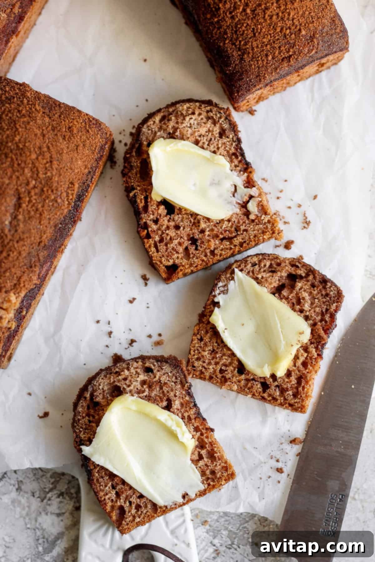 Two slices of dark bread with butter on a parchment paper, next to a knife with a partially visible bread loaf, showcasing the cinnamon crunch topping.