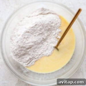 A clear bowl contains a mixture of liquid ingredients and dry flour with a gold spoon, set on a textured white surface, showing the wet and dry ingredients combined.