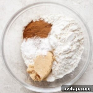 A glass bowl containing flour, brown sugar, cinnamon, baking powder, and baking soda, on a light textured surface, ready to be mixed.