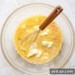 A glass bowl containing a beaten egg mixture, maple syrup, vanilla extract, almond milk, avocado oil, and Greek yogurt, with a whisk resting inside.