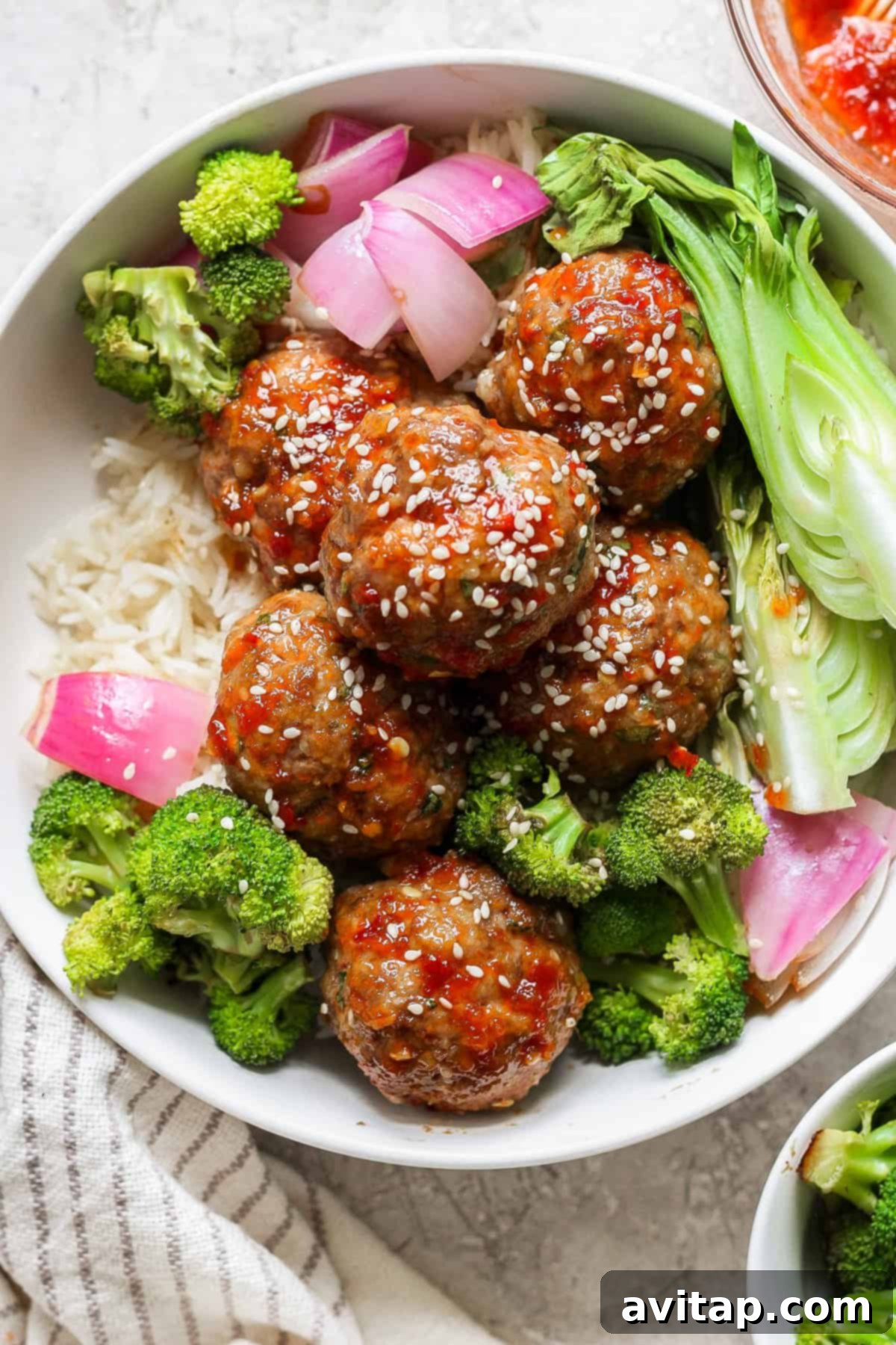 A vibrant bowl featuring glazed sweet chili garlic meatballs, garnished with sesame seeds, accompanied by tender roasted broccoli, bok choy, and sliced red onion, all served over fluffy rice.
