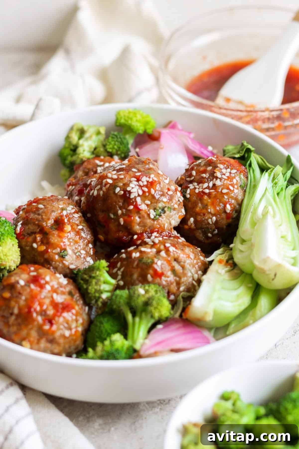 A beautifully plated dish of sweet chili garlic meatballs, glistening with sauce and topped with sesame seeds, alongside roasted broccoli, bok choy, and red onions. A small bowl of extra sauce and a basting brush are in the background.
