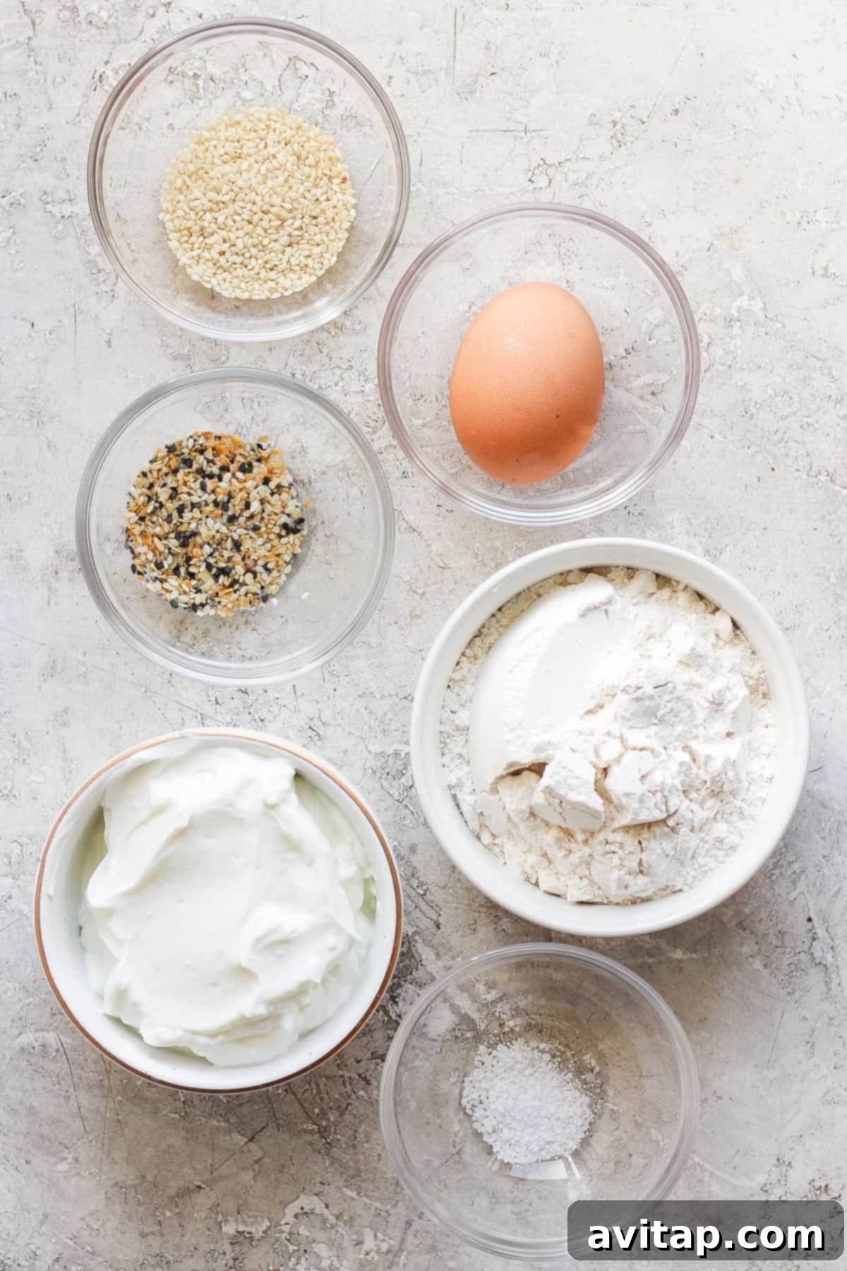 Six small bowls arranged on a textured surface, holding various bagel ingredients including sesame seeds, spices, flour, creamy yogurt, and salt, with one egg.