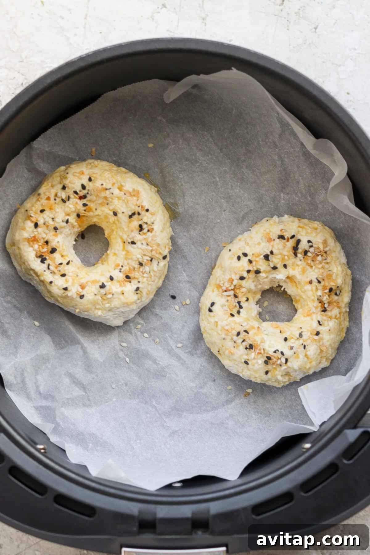 Two bagels with seasoning on top are cooking on parchment paper inside an air fryer basket, showing the baking process.