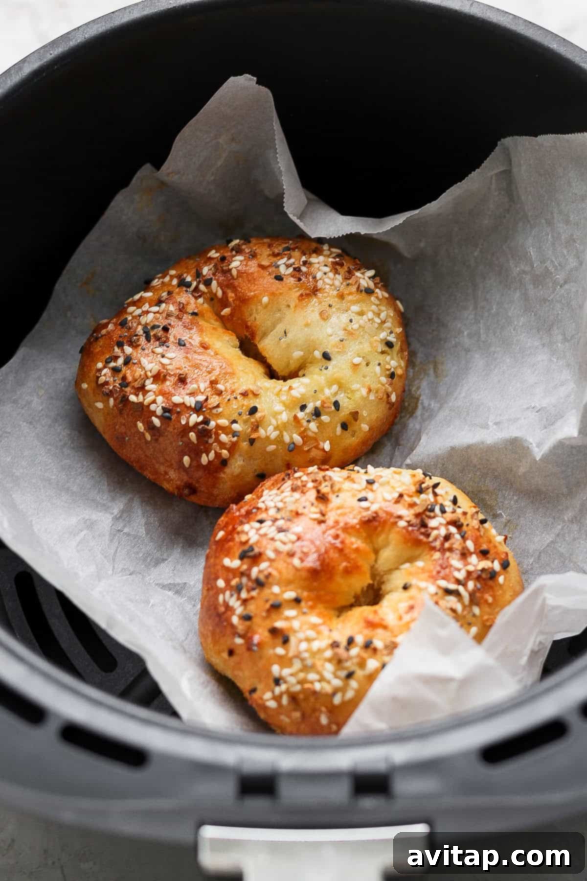 Two golden-brown bagels, generously topped with sesame and poppy seeds, baking on parchment paper inside an air fryer basket.