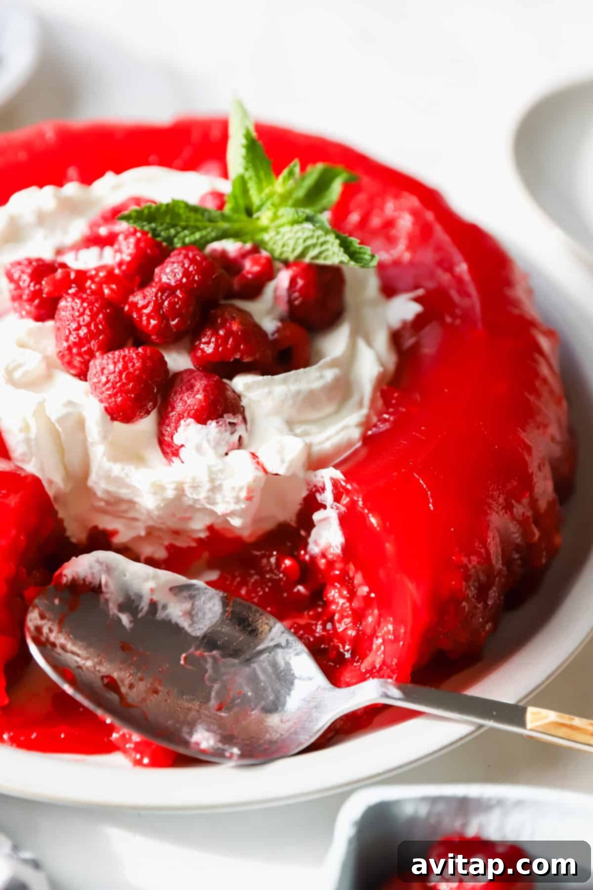 A beautifully set red gelatin dessert, generously topped with fluffy whipped cream, fresh, glistening raspberries, and a sprig of fragrant mint. A spoon rests gracefully on the white plate beside this inviting treat.