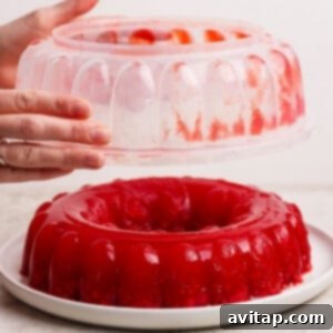 Hands demonstrating the removal of a jello mold from a red gelatin dessert, which now stands perfectly formed on a white serving plate.