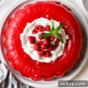 A vibrant red gelatin dessert crowned with fluffy whipped cream, fresh raspberries, and a sprig of mint on a white plate, presented alongside a striped cloth and a small dish of extra raspberries, ready for serving.