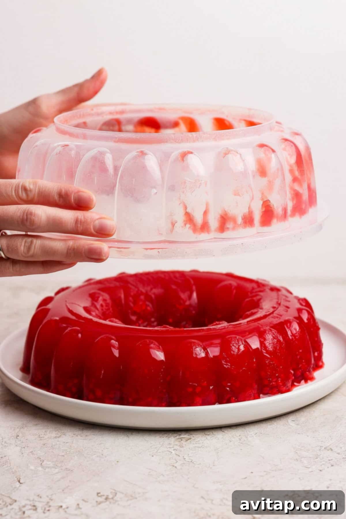 A pair of hands carefully lifting a jello mold away from a vibrant red gelatin dessert, which now rests perfectly formed on a pristine white plate.