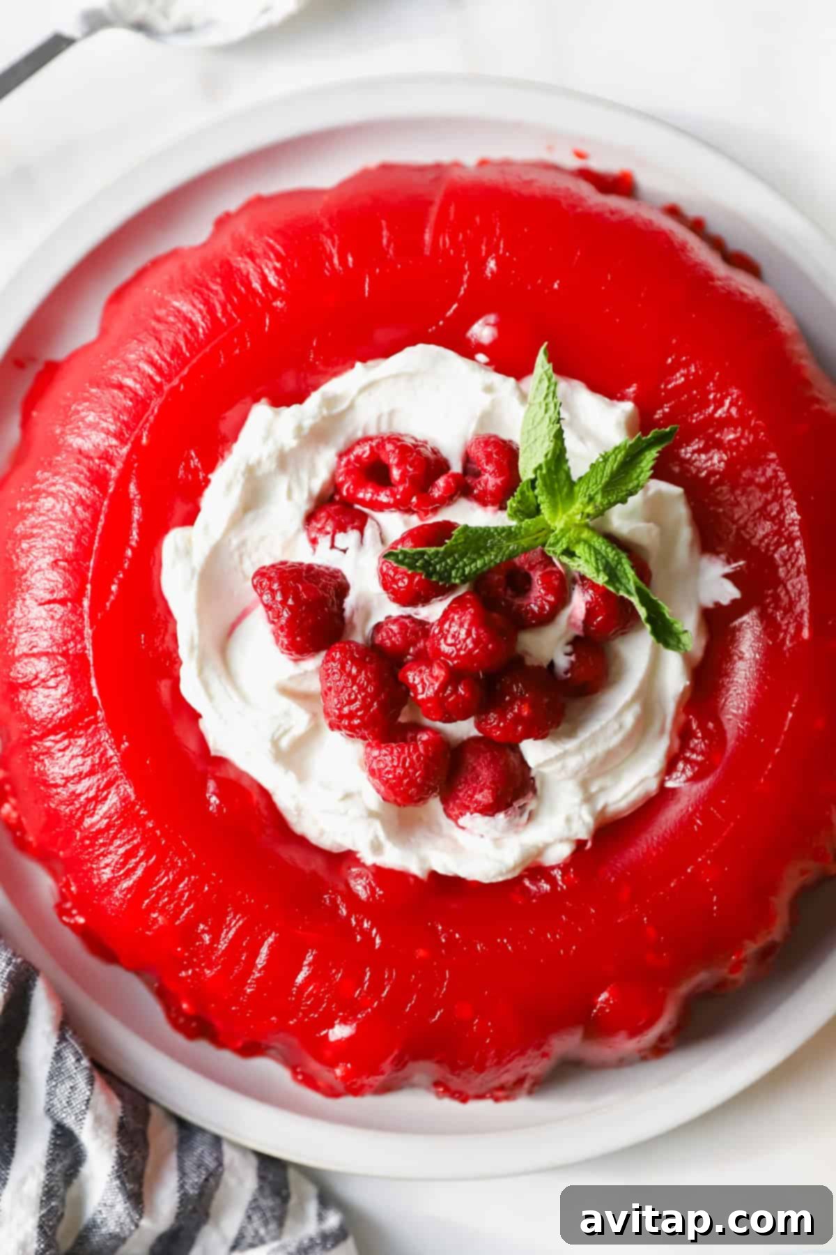 A vibrant red gelatin mold, elegantly presented on a white plate, crowned with a generous swirl of whipped cream, fresh raspberries, and a delicate sprig of mint.