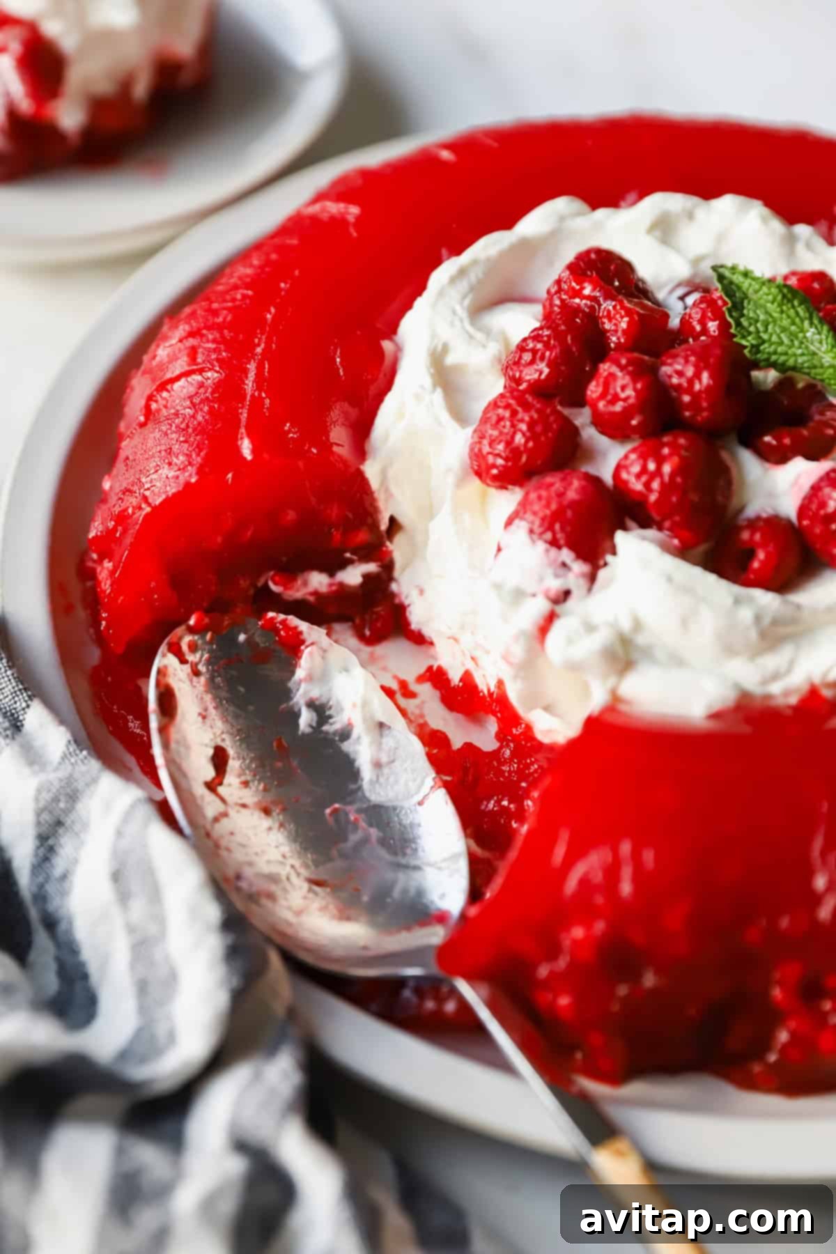 A vibrant red gelatin dessert, elegantly served on a white plate, adorned with a swirl of whipped cream and fresh raspberries. A portion has been removed, and a spoon rests invitingly nearby, hinting at its deliciousness.
