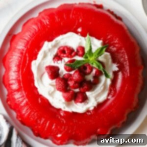 A red gelatin dessert topped with whipped cream, fresh raspberries, and mint leaves on a white plate, ready to be served.