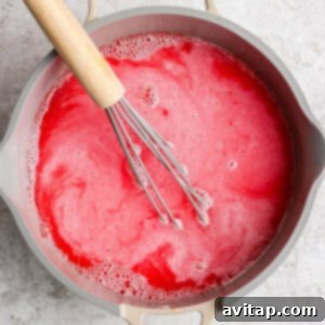A whisk in a pot of frothy, red liquid on a light-colored surface, illustrating the initial mixing step for the jello mold recipe.