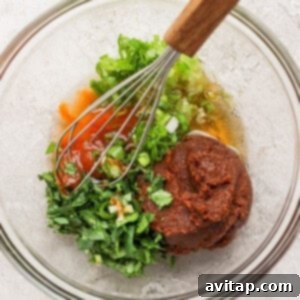 A glass bowl containing chopped green herbs, red sauce, a thick brown paste, and a whisk with a wooden handle on a light-colored surface, illustrating marinade preparation.