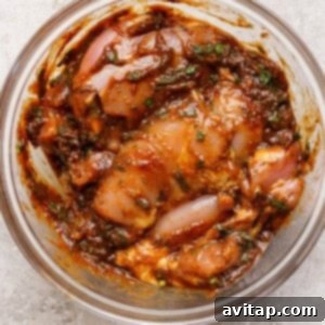 A clear glass bowl containing marinated pieces of raw chicken mixed with herbs and spices in a thick, reddish-brown sauce, ready for refrigeration.