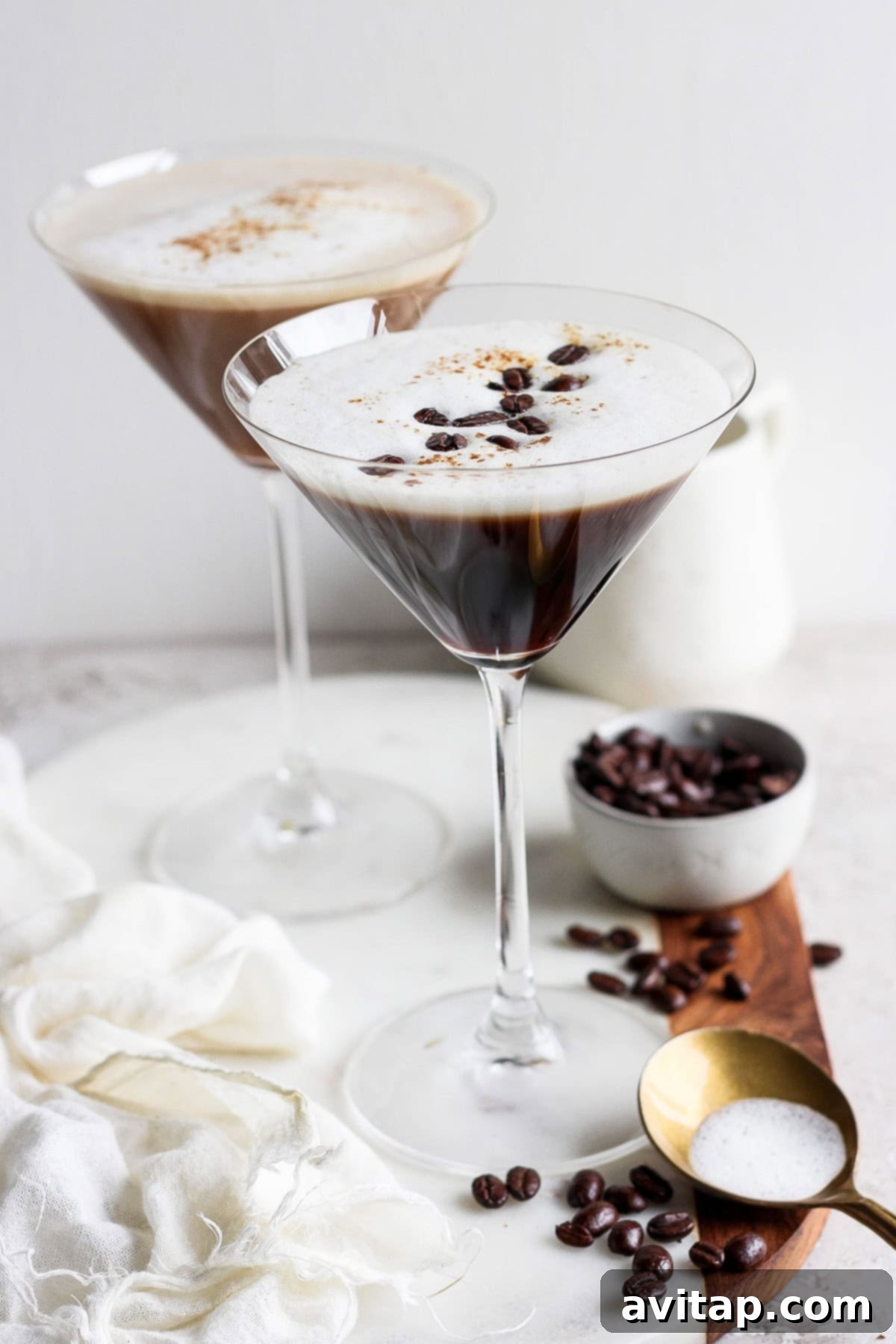 Velvet Brown Sugar Espresso Martini 2 Two espresso martinis in martini glasses, garnished with coffee beans, on a wooden board with a small bowl of coffee beans and a brass spoon nearby. A white cloth is draped on the left.