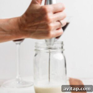A hand holds a milk frother in a glass jar on a marble board, creating foam. Two stem glasses are in the background.