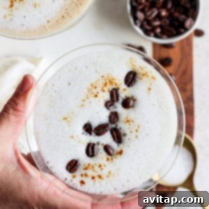 A hand holding a frothy coffee drink in a glass, garnished with coffee beans and cinnamon. Another glass and a bowl of coffee beans are visible in the background.