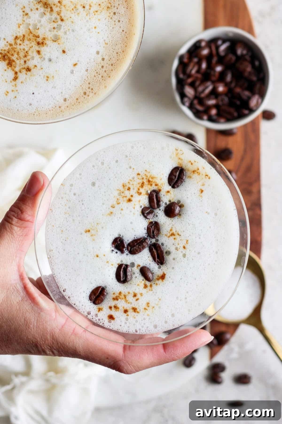 Velvet Brown Sugar Espresso Martini 5 A hand holding a frothy coffee drink in a glass, garnished with coffee beans and cinnamon. Another glass and a bowl of coffee beans are visible in the background.