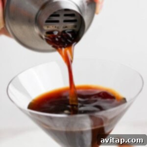 A hand pouring a dark liquid from a metal shaker into a martini glass.