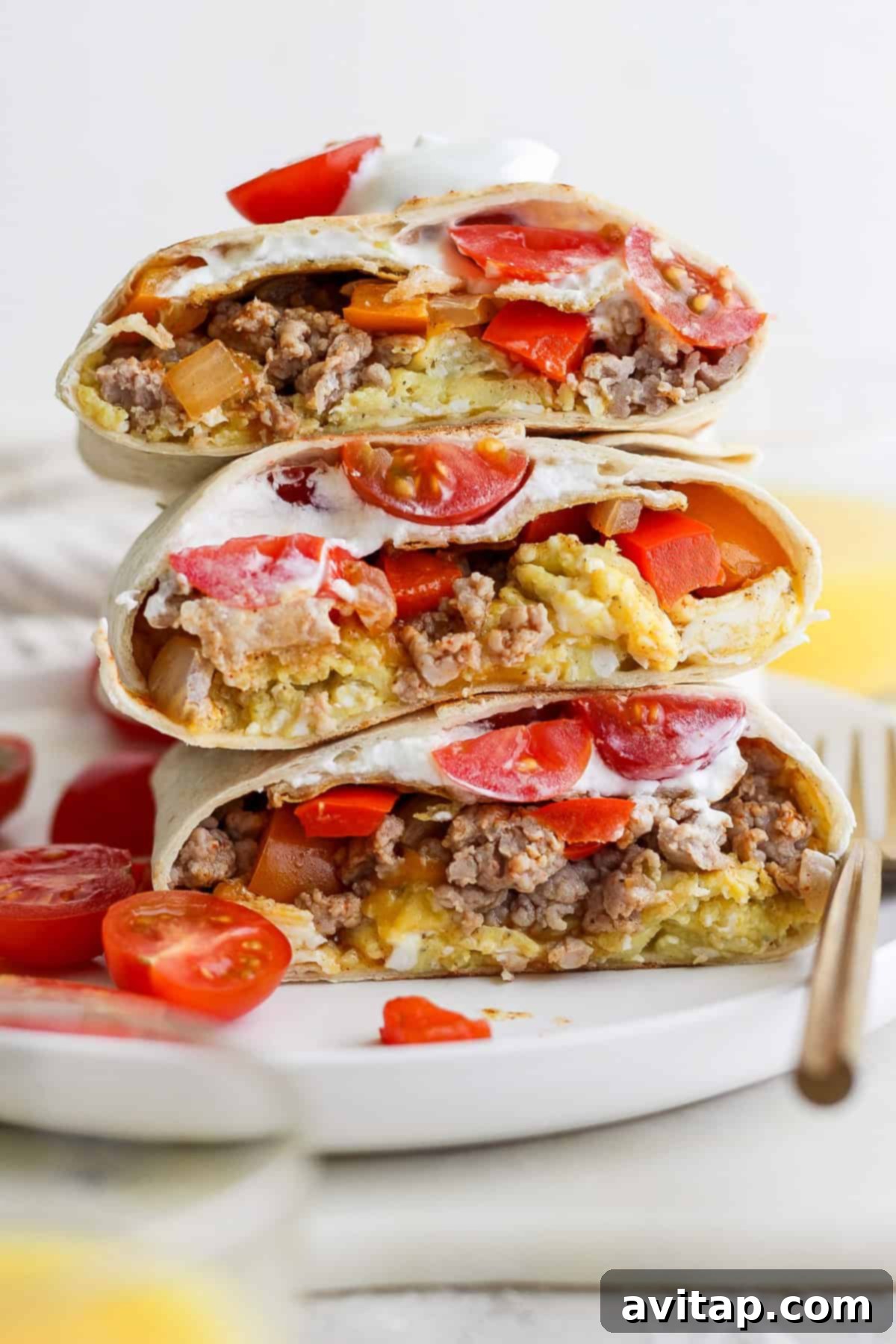 Stacked burritos filled with ground meat, cheese, tomatoes, and sour cream on a white plate. Sliced tomatoes are scattered around.