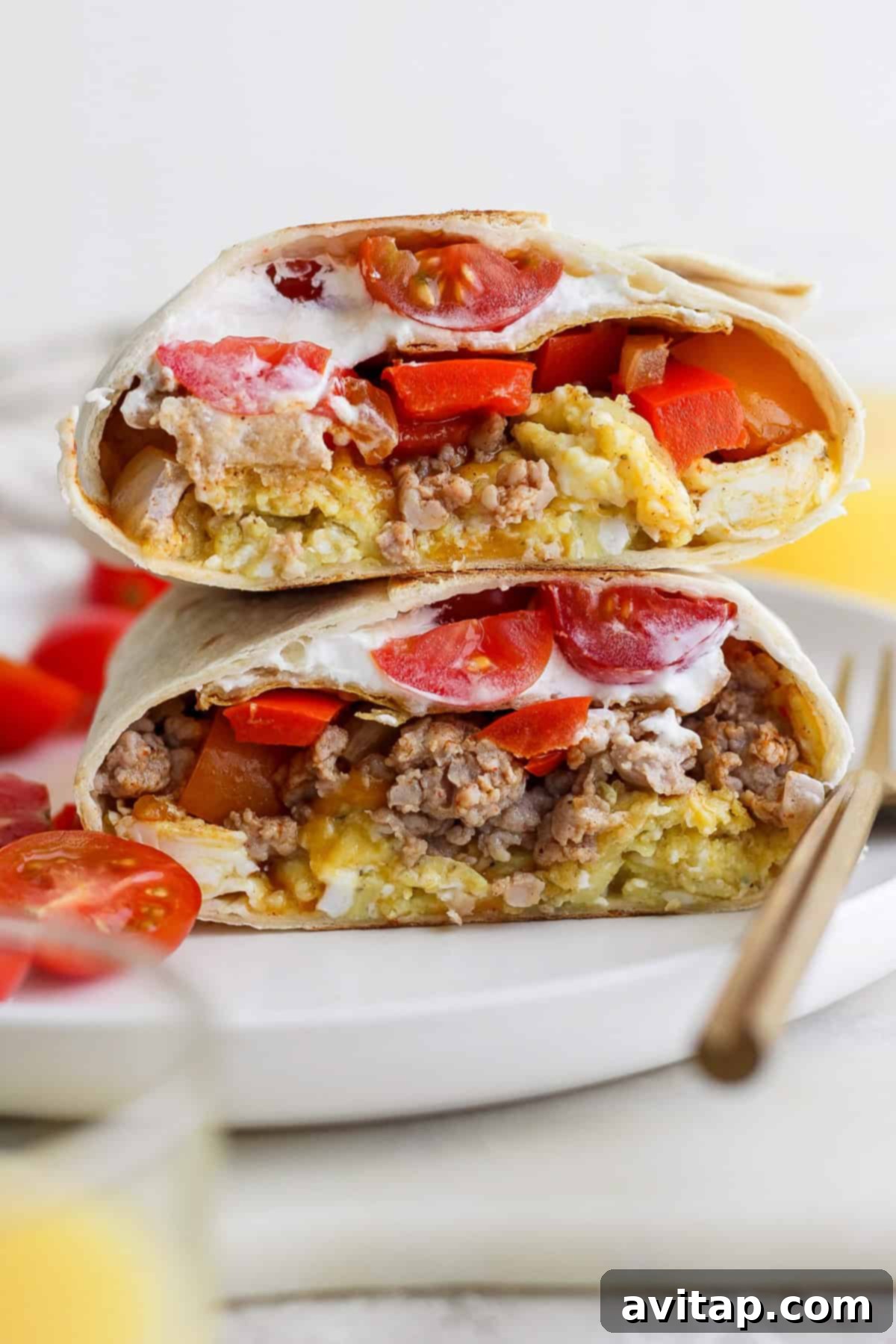 Two halves of a breakfast burrito filled with eggs, sausage, tomatoes, and topped with sour cream are stacked on a white plate.