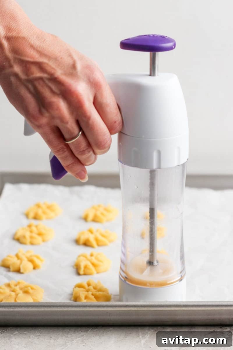 Mabel's Classic Spritz Cookies 4 A hand pressing cookie dough onto a baking sheet using a cookie press, forming flower-shaped cookies.