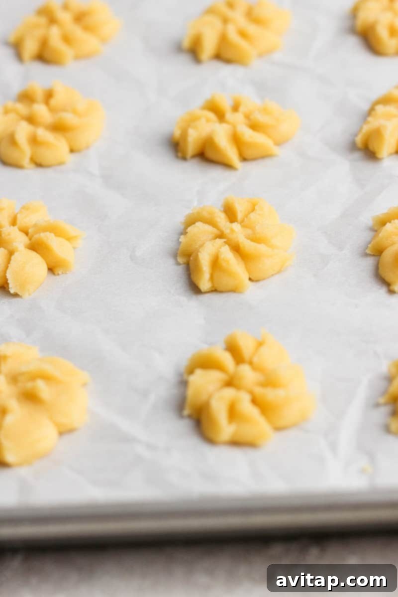 Mabel's Classic Spritz Cookies 5 Raw cookie dough shaped into floral patterns on a parchment-lined baking sheet, ready for baking.
