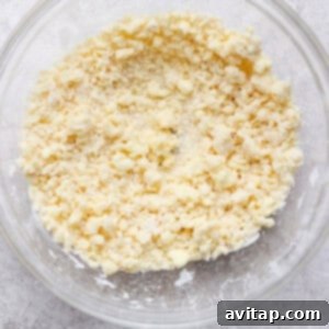 A clear glass bowl containing a crumbly mixture of flour and butter, sitting on a textured gray surface.