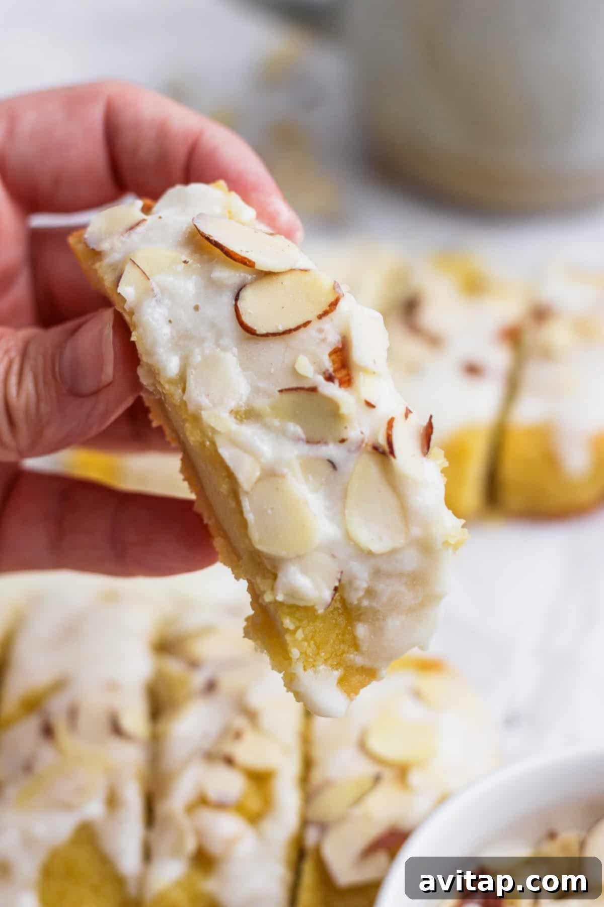 A hand holding a slice of almond-topped dessert bar with white icing. Other similar dessert bars are visible in the background.