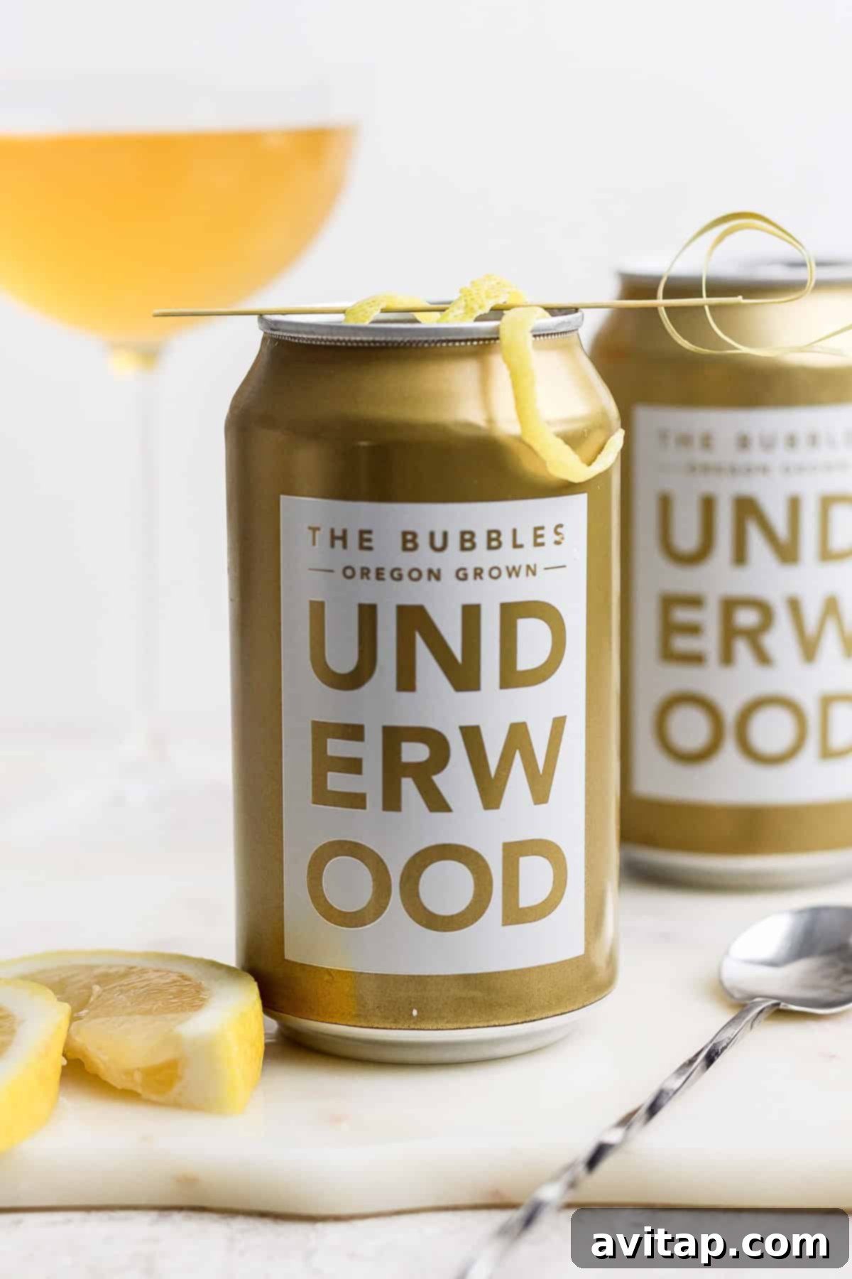 Two cans of Underwood "The Bubbles" canned champagne cocktail with lemon garnish grace the table, accompanied by a spoon and lemon slices. A wine glass adds elegance in the background.