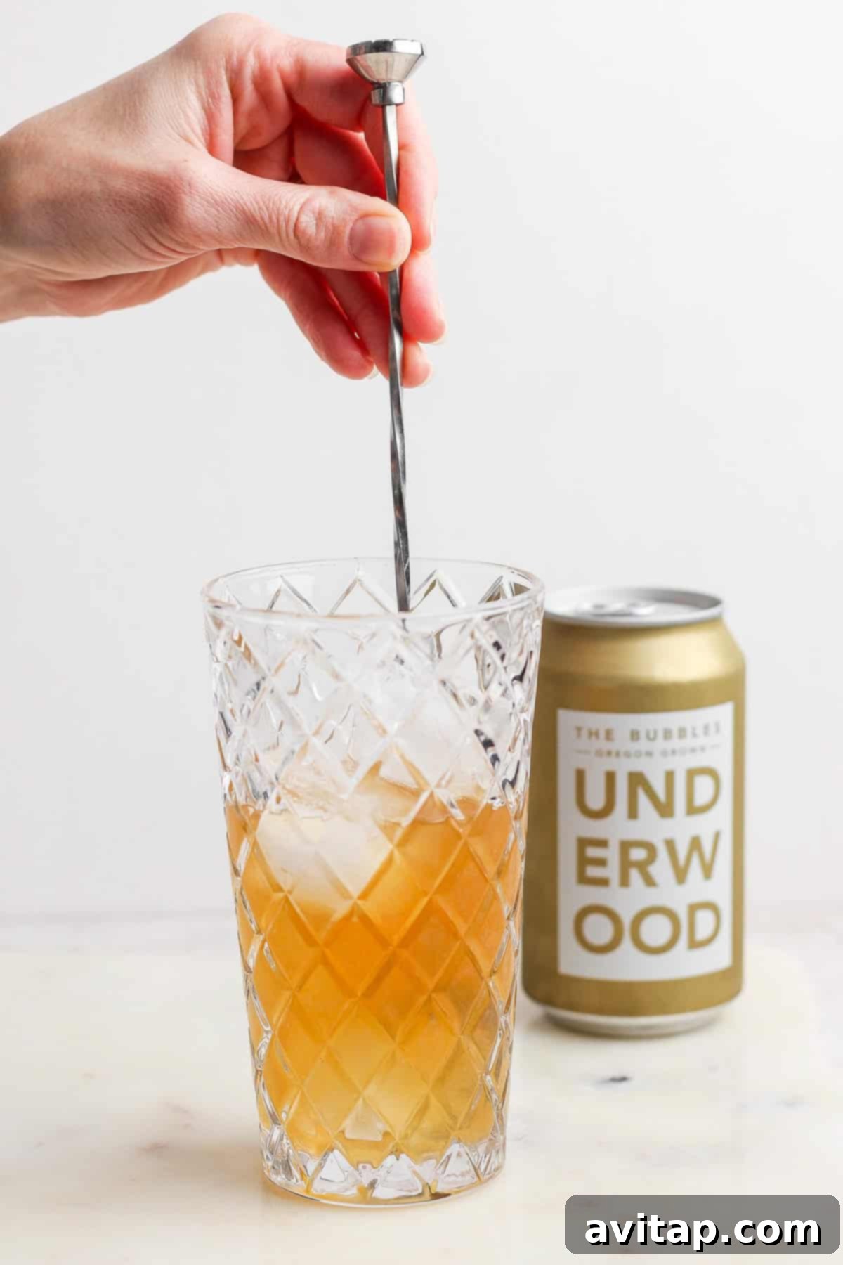 A hand stirs a canned champagne cocktail with ice in a diamond-patterned glass, while an unopened can of Underwood sparkling wine sits elegantly in the background.