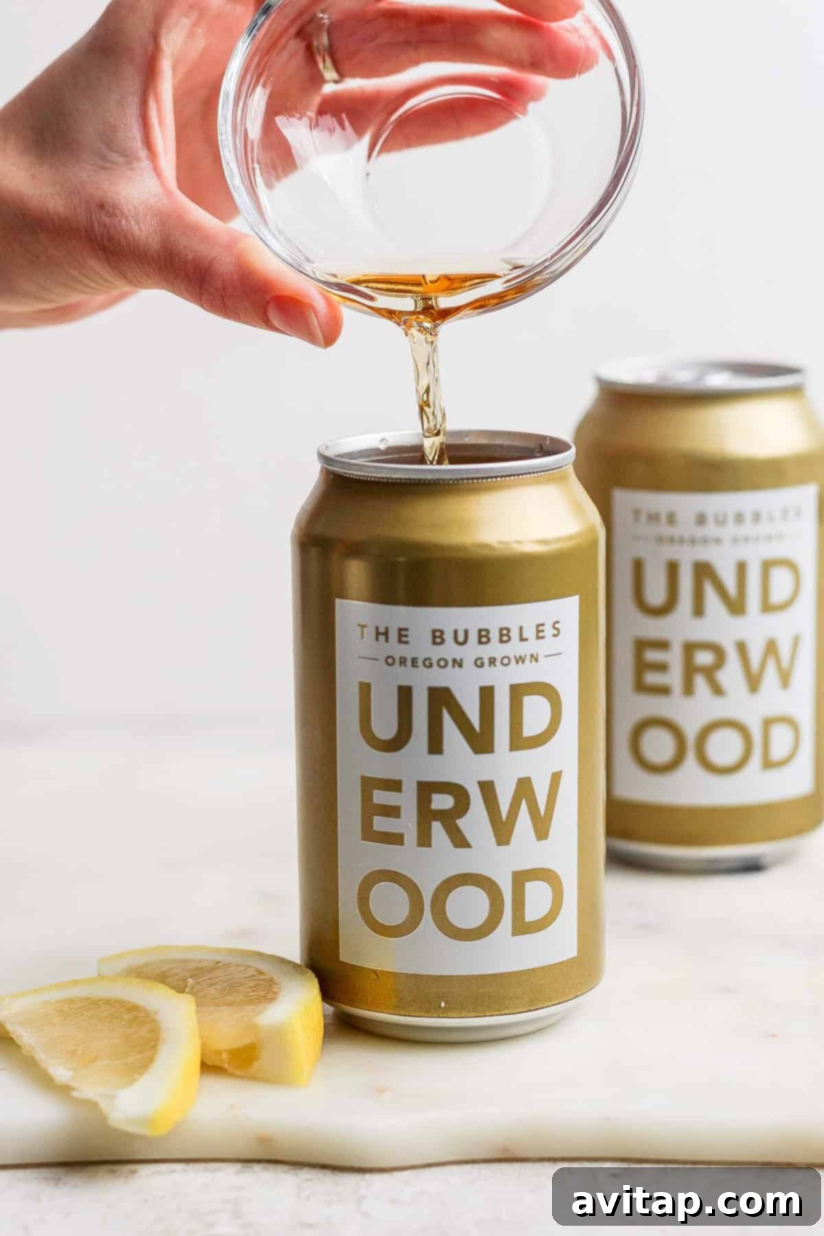 A hand elegantly pours liquid from a glass into a can labeled "Underwood: The Bubbles Oregon Grown," creating the perfect canned champagne cocktail. Two lemon slices and another can are subtly visible in the background, adding a touch of zest to this sparkling scene.