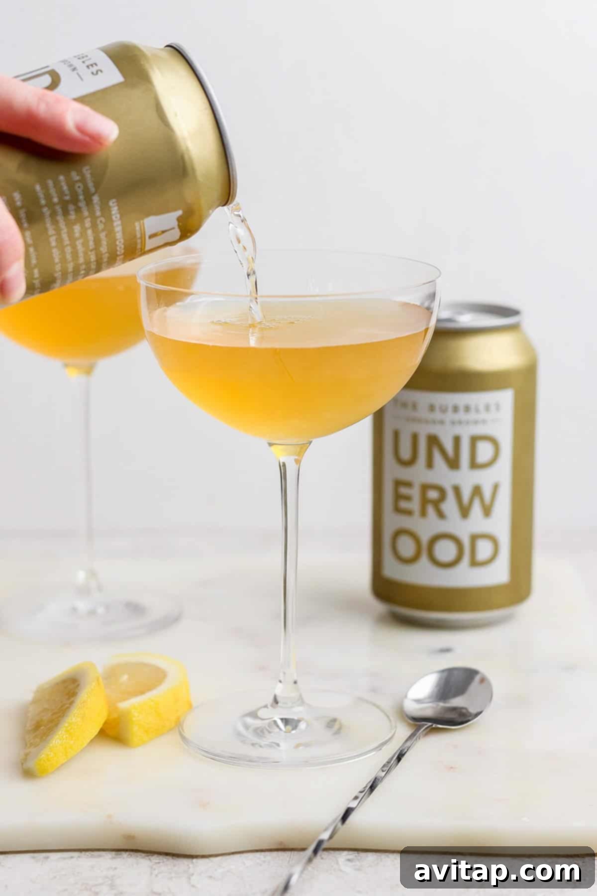 A can labeled "Underwood" is pouring a golden liquid, reminiscent of a canned champagne cocktail, into a coupe glass. Another filled glass, the can itself, lemon wedges, and a spoon are arranged on the marble surface.