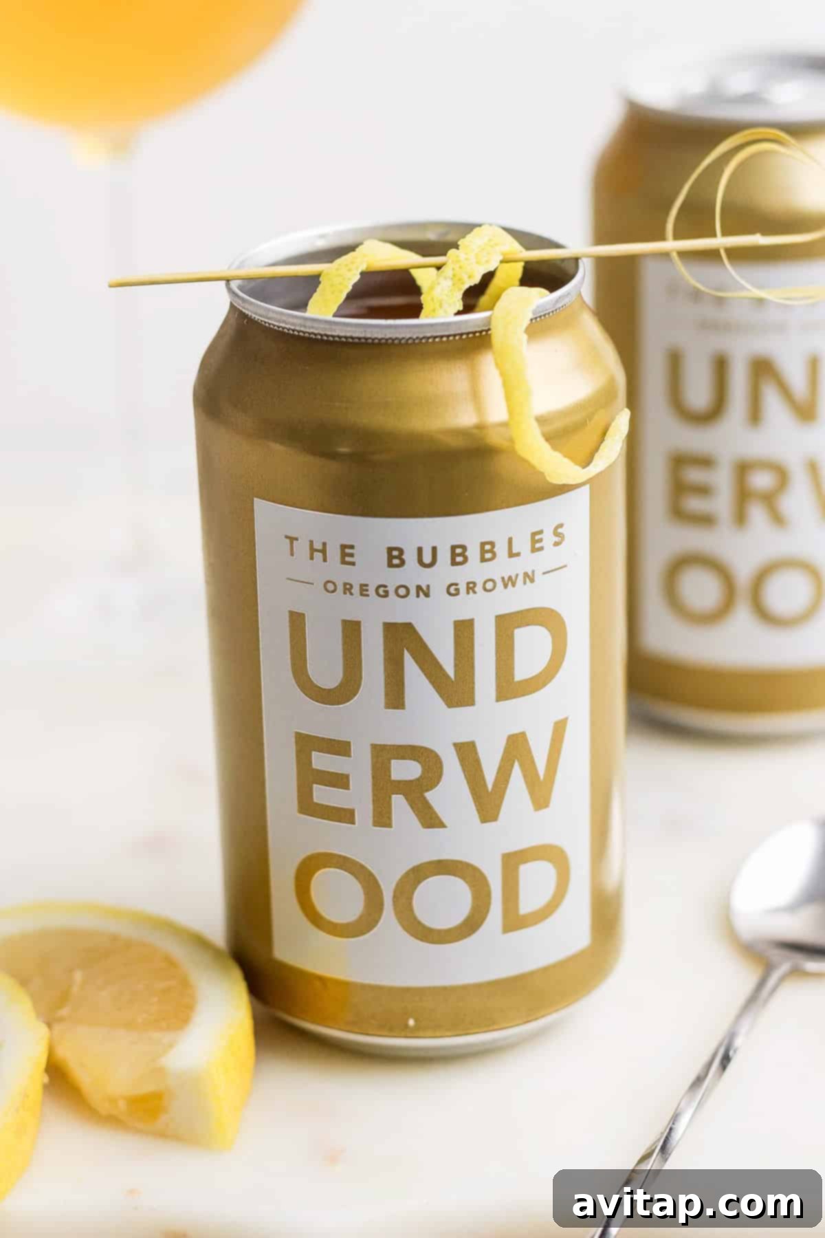 Underwood sparkling wine can with a lemon twist garnish sits next to lemon slices and a spoon on a white surface, creating an enticing canned champagne cocktail experience.