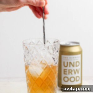 A hand stirs a canned champagne cocktail with ice in a diamond-patterned glass, while an unopened can of Underwood sparkling wine sits elegantly in the background.
