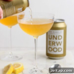 A can labeled "Underwood" is pouring a golden liquid, reminiscent of a canned champagne cocktail, into a coupe glass. Another filled glass, the can itself, lemon wedges, and a spoon are arranged on the marble surface.