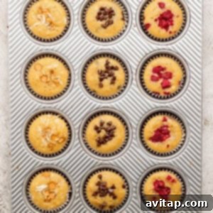 A muffin tray featuring 12 freshly baked muffins, some topped with slivered almonds, others with chocolate chips, and bright red raspberries, cooling after baking.