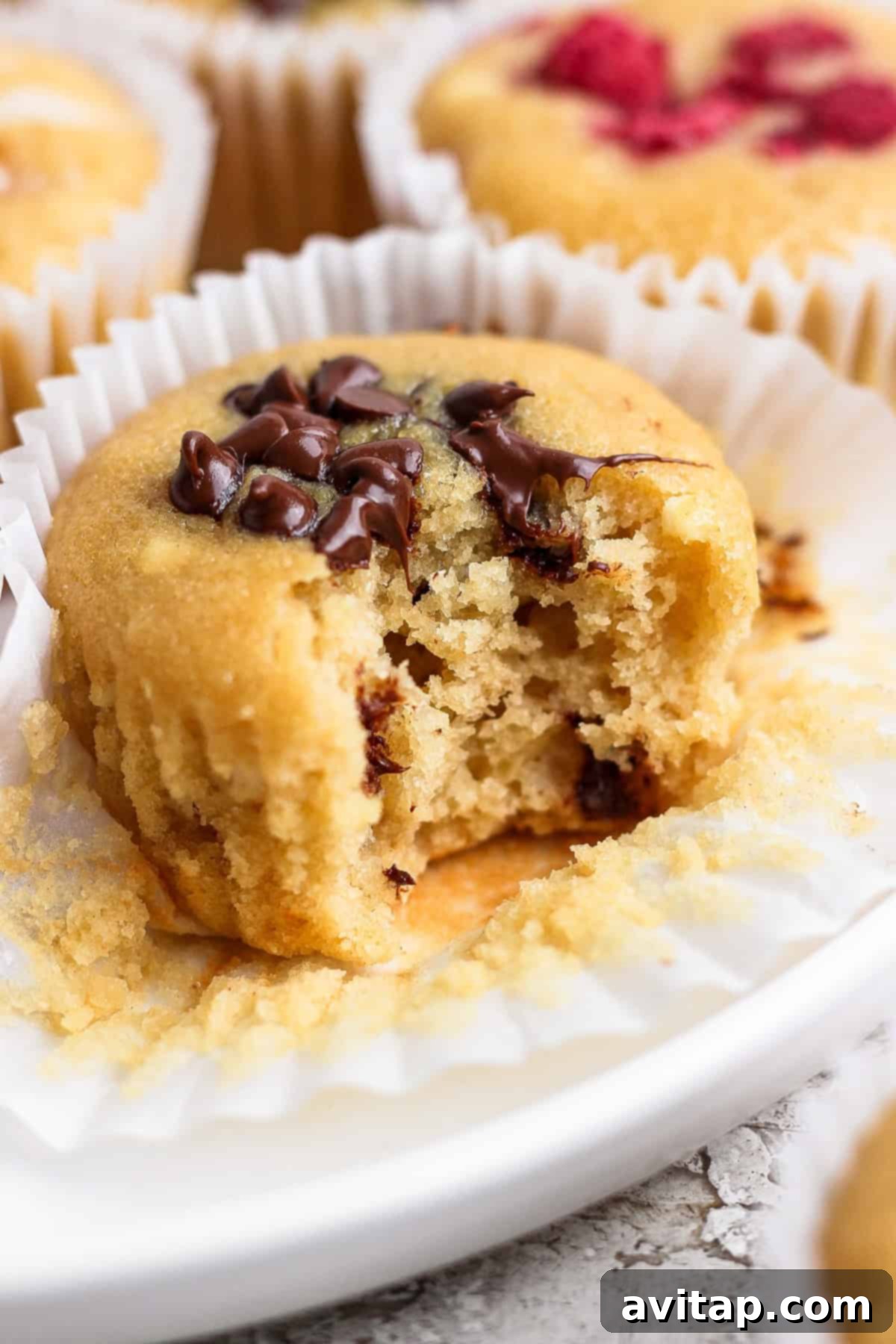 A close-up of a partially eaten, golden-brown muffin adorned with melted chocolate chips, resting invitingly in a white paper liner on a light surface, highlighting its moist and tender interior.