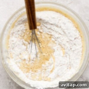 A glass bowl with a wooden-handled whisk mixing flour into a wet batter mixture on a textured kitchen counter.