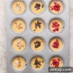 Muffin batter already portioned into a muffin tray, with various toppings like coconut, chocolate chips, and raspberries visible on top before baking.