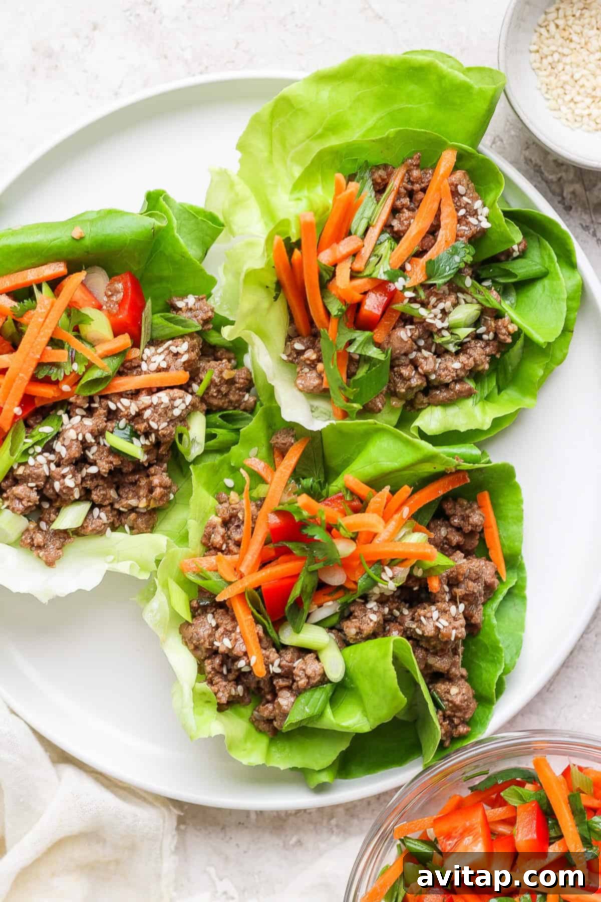 Four lettuce wraps filled with seasoned ground beef, shredded carrots, diced red bell peppers, and fresh green onions, elegantly arranged on a clean white plate.
