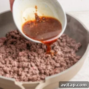A hand pours a brown sauce from a white bowl onto cooked ground beef in a beige skillet.