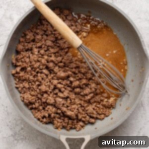 Ground beef cooking in a skillet with a whisk resting on the side and a pool of juices visible.