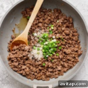 Ground beef cooked in a pan with a wooden spoon, topped with sesame seeds and chopped green onions.