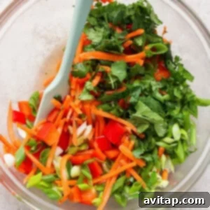 A glass bowl filled with chopped vegetables, including red bell peppers, carrots, green onions, cilantro, and green bell peppers, with a light blue spatula.