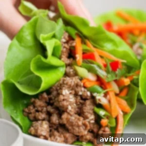 A hand holding a lettuce wrap filled with ground meat, shredded carrots, and herbs on a white plate.