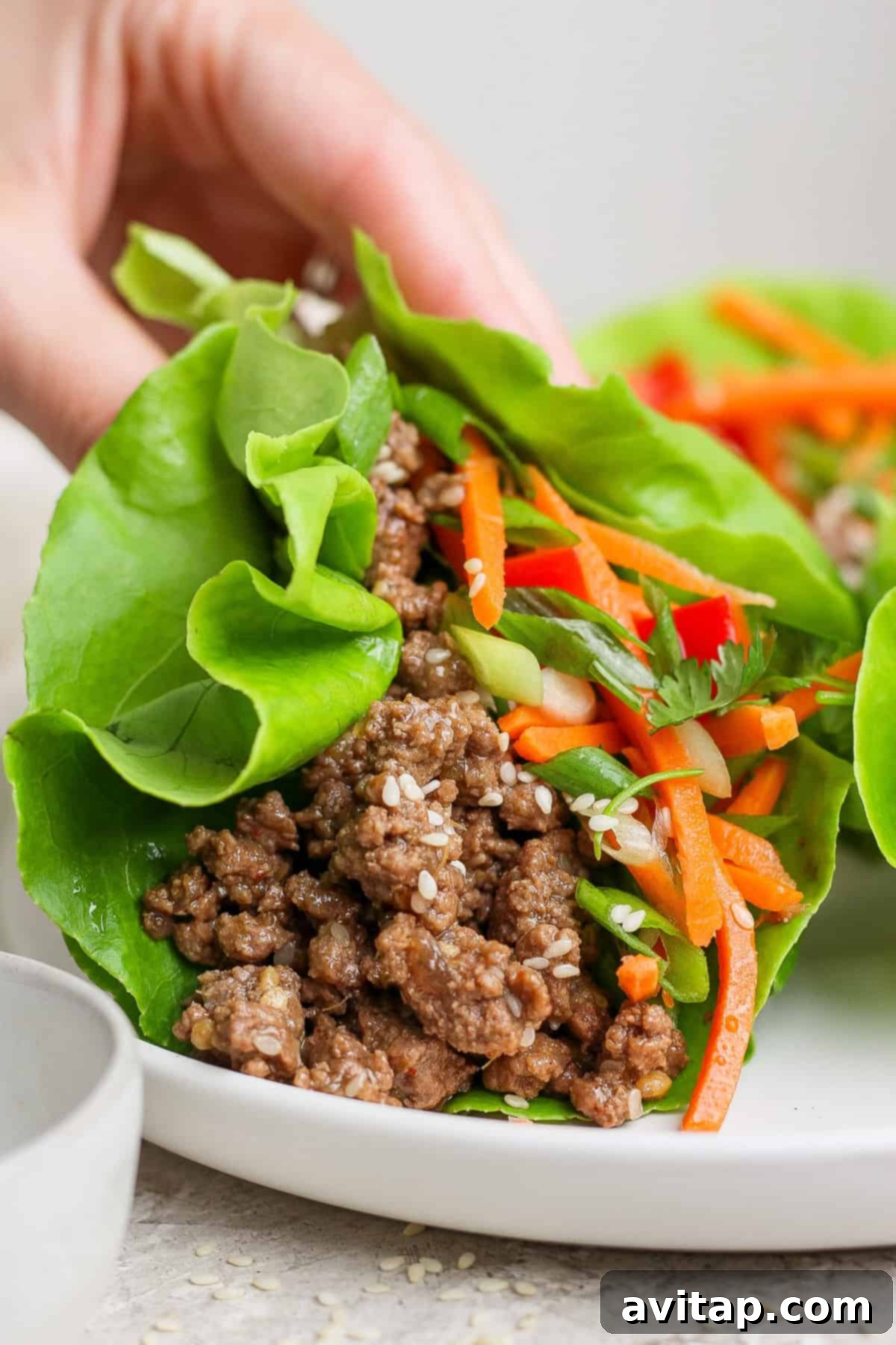A hand gently holds a perfectly assembled Korean Beef Lettuce Wrap, showcasing its rich filling of ground meat, vibrant shredded carrots, and fresh herbs. The wrap is presented on a pristine white plate, inviting a delicious bite.