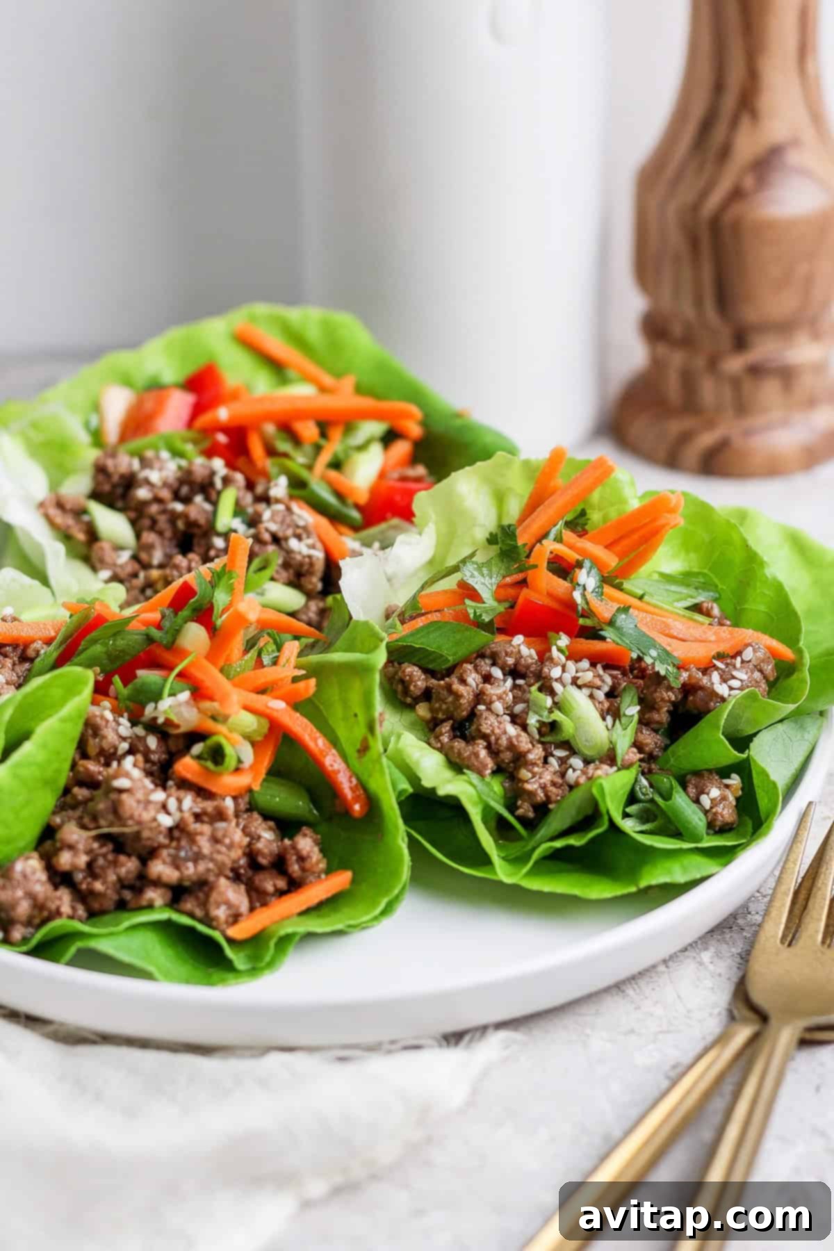 Freshly prepared lettuce wraps filled with seasoned minced meat, thinly sliced carrots, a sprinkle of sesame seeds, and chopped green onions are artfully arranged on a pristine white plate. Elegant gold cutlery is visible in the foreground, suggesting a gourmet dining experience.